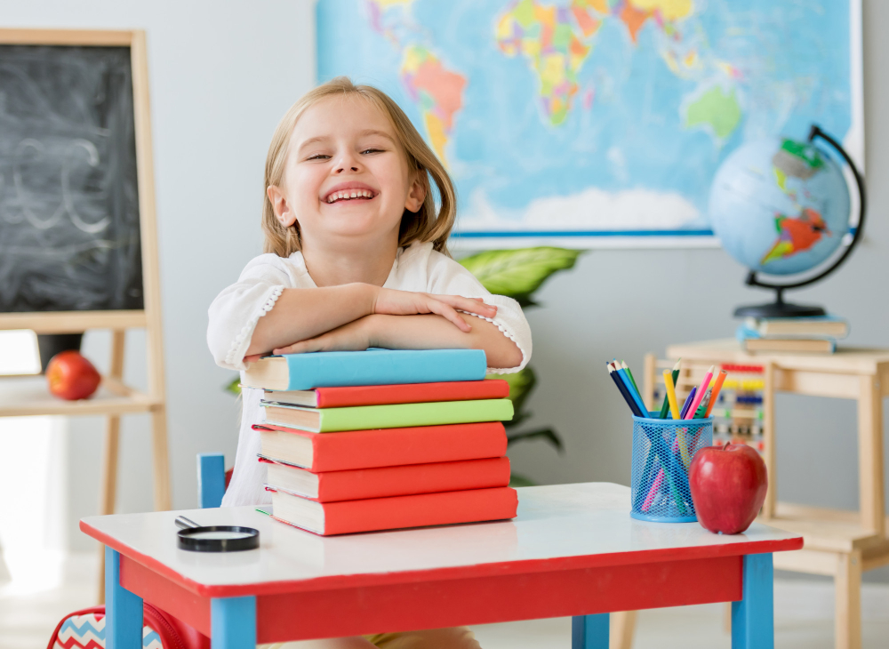 little smiling blond girl sitting white desk holding hands books spacious school class